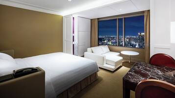 Ein Hotelzimmer mit Bett, einer Couch, einem Esstisch und Blick auf eine nächtliche Stadtansicht.