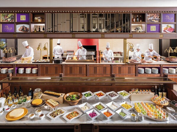 Una cocina profesional con chefs preparando alimentos, un bufé con diversos platos y bebidas, y una pared decorativa con fotografías enmarcadas.