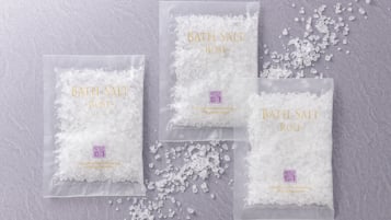 Drei Packungen Badesalz mit der Aufschrift 'Badesalz ~ Rose ~' und einem violetten Logo, platziert auf einer strukturierten Oberfläche.