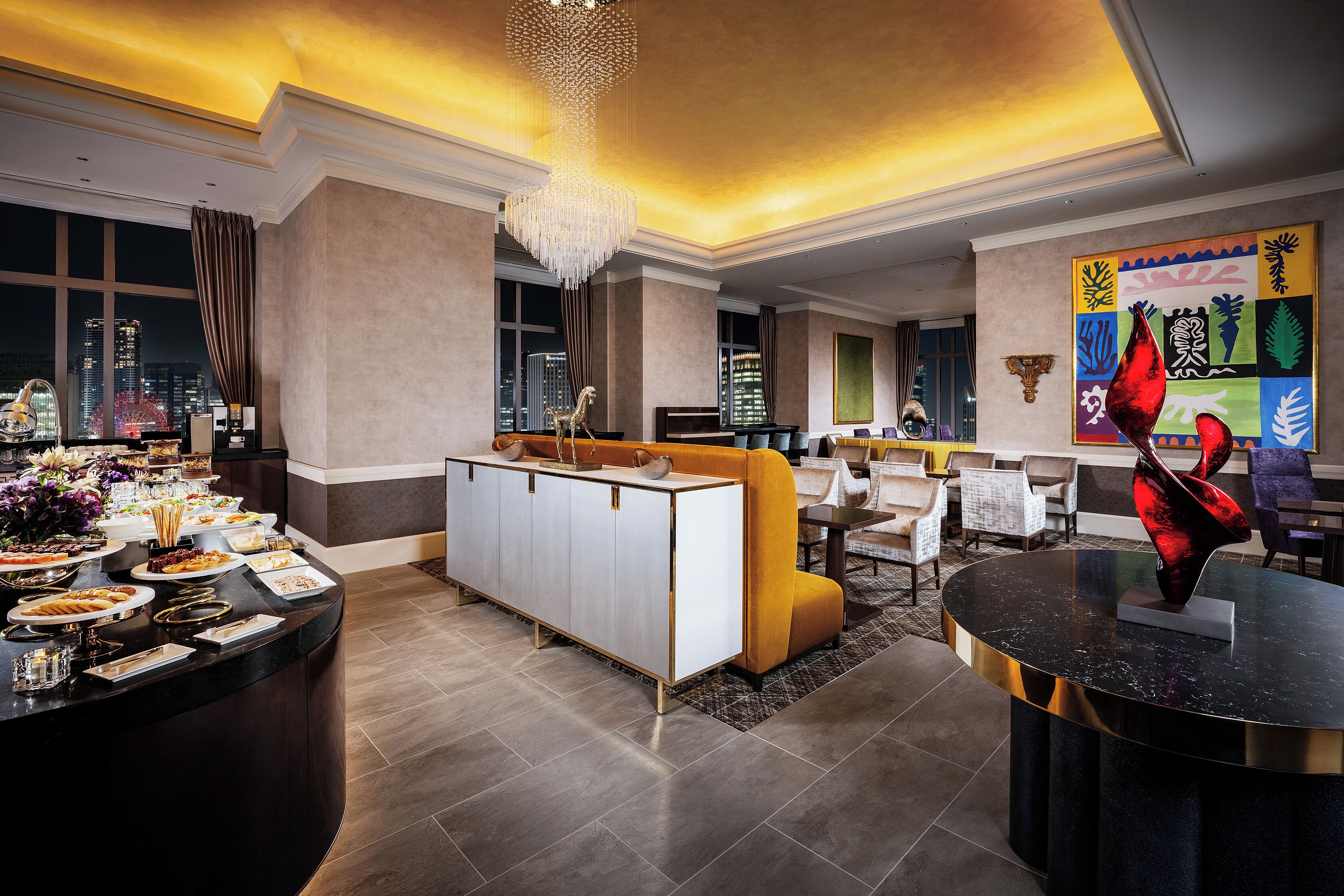Eine moderne Hotelhalle mit einer gebogenen Rezeption, einer Sitzecke mit gelben Stühlen, einem Chandelier und einem Blick auf die Stadt durch große Fenster.