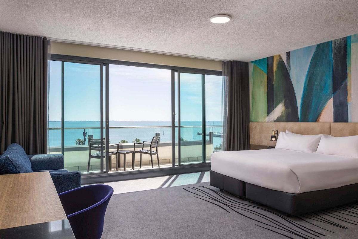 Habitación de hotel con una cama grande, un escritorio, una silla, un balcón con vistas al mar y un cuadro abstracto moderno en la pared.