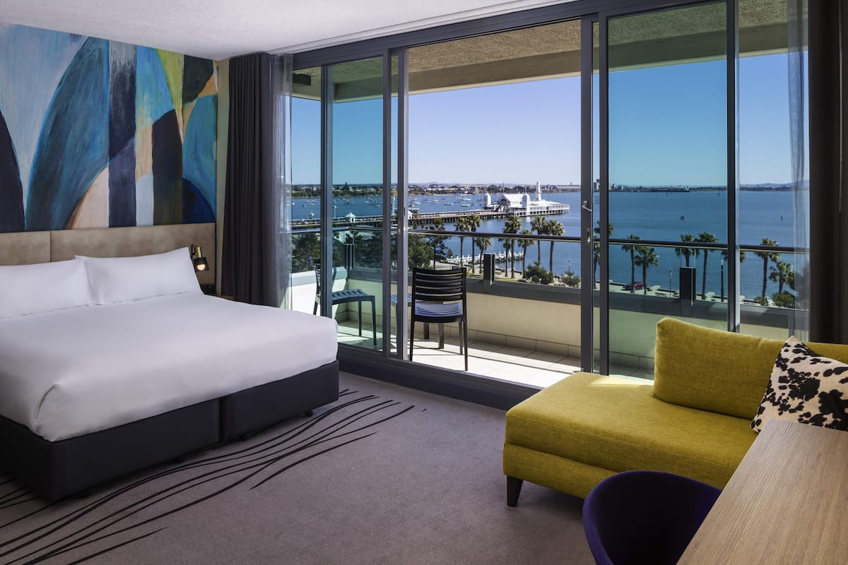 Habitación de hotel con cama, una mesita, una silla y un sofá. La habitación tiene ventanas amplias con vistas a un puerto deportivo y al mar.