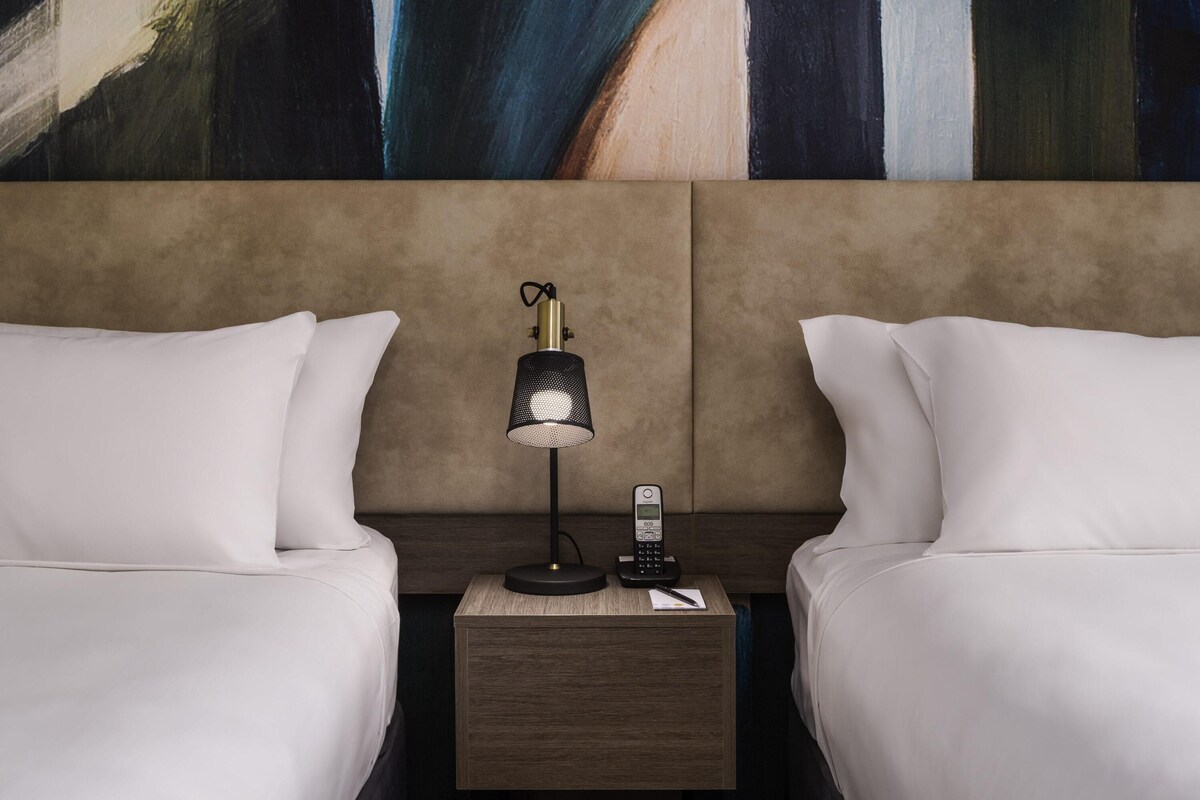 A hotel room with two beds, a nightstand with a lamp, a phone, and a painting on the wall.