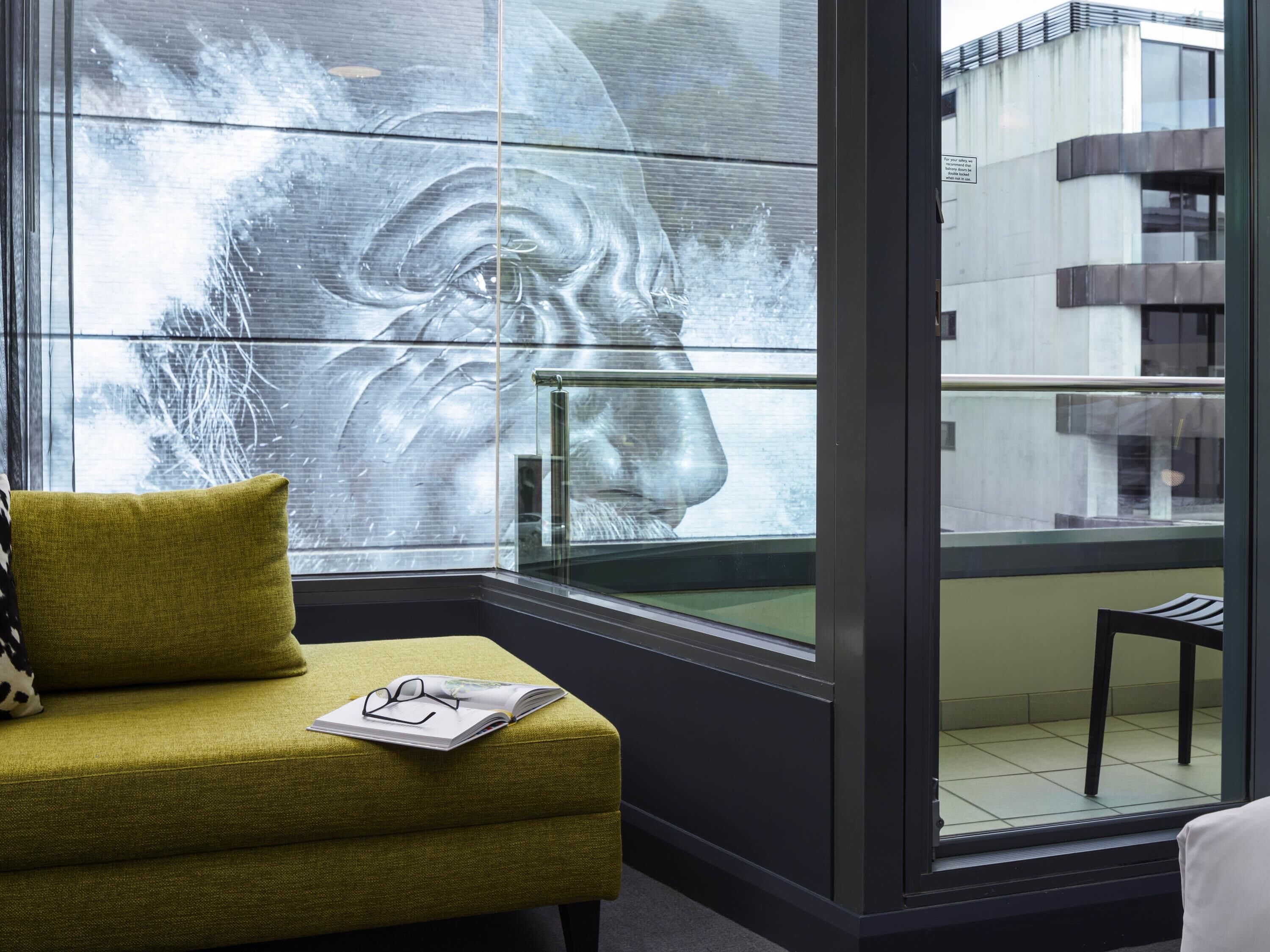 Ein modernes Zimmer mit einem grünen Sofa, einem Glastisch mit einer Zeitschrift und Gläsern sowie einem großen Fenster mit unscharfem Hintergrund.