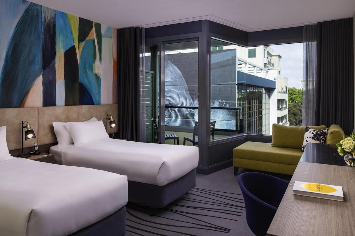 Habitación de hotel con dos camas, un escritorio, una silla y un amplio ventanal con vista a la ciudad.