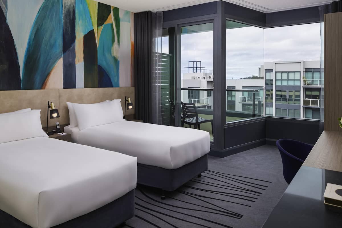 Una habitación de hotel moderna con dos camas, un ventanal con vistas a la ciudad y un cuadro abstracto contemporáneo en la pared.