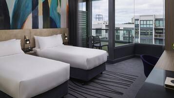 A modern hotel room with two beds, a large window with a city view, and a contemporary abstract painting on the wall.