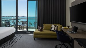 Una habitación de hotel con cama, sofá, escritorio con computadora portátil, televisión y vista al océano y palmeras.