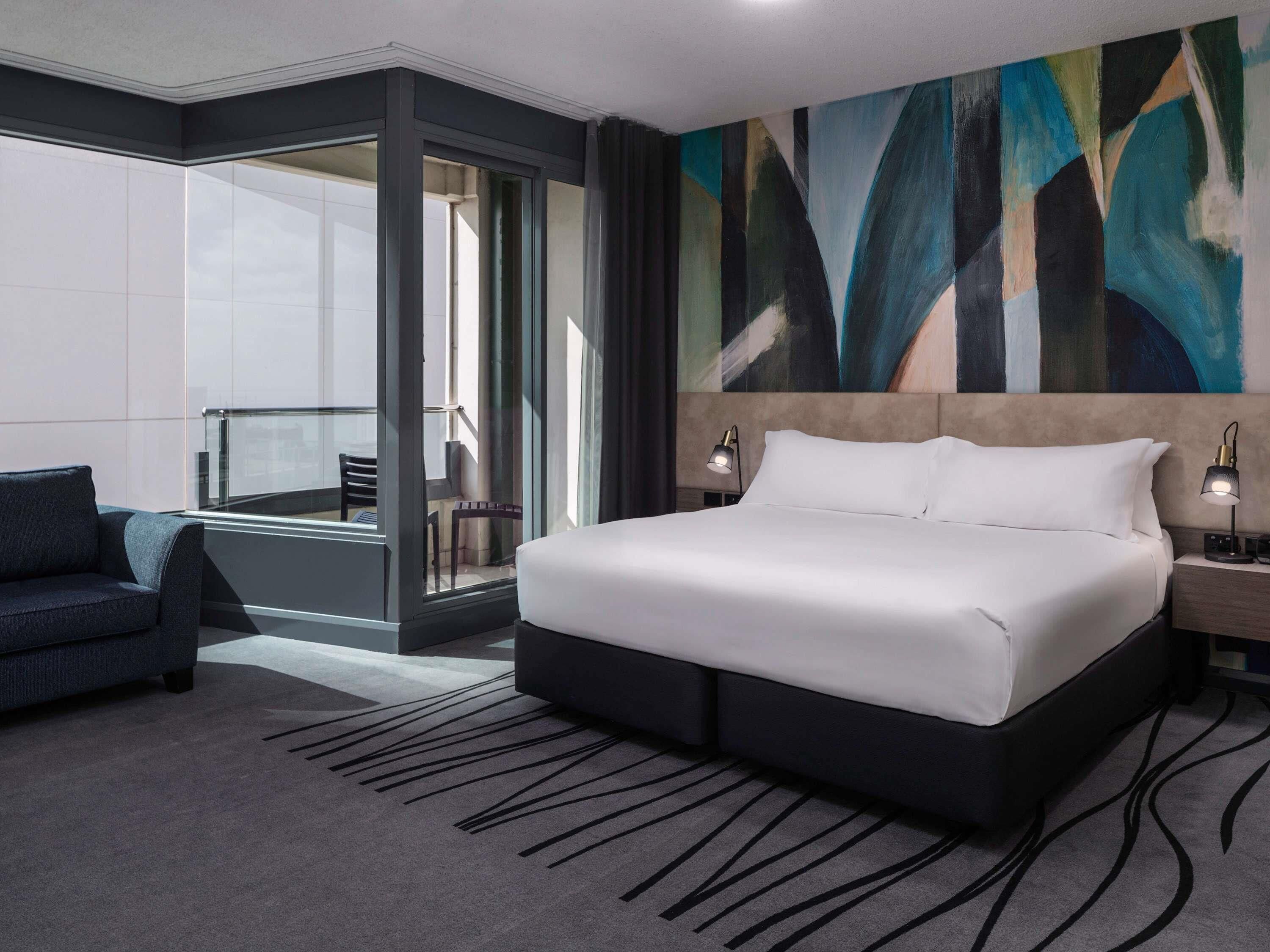 A modern hotel room with a large bed, a seating area with a chair, a glass partition, and a large abstract painting on the wall.