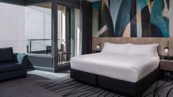 A modern hotel room with a large bed, a seating area with a chair, a glass partition, and a large abstract painting on the wall.