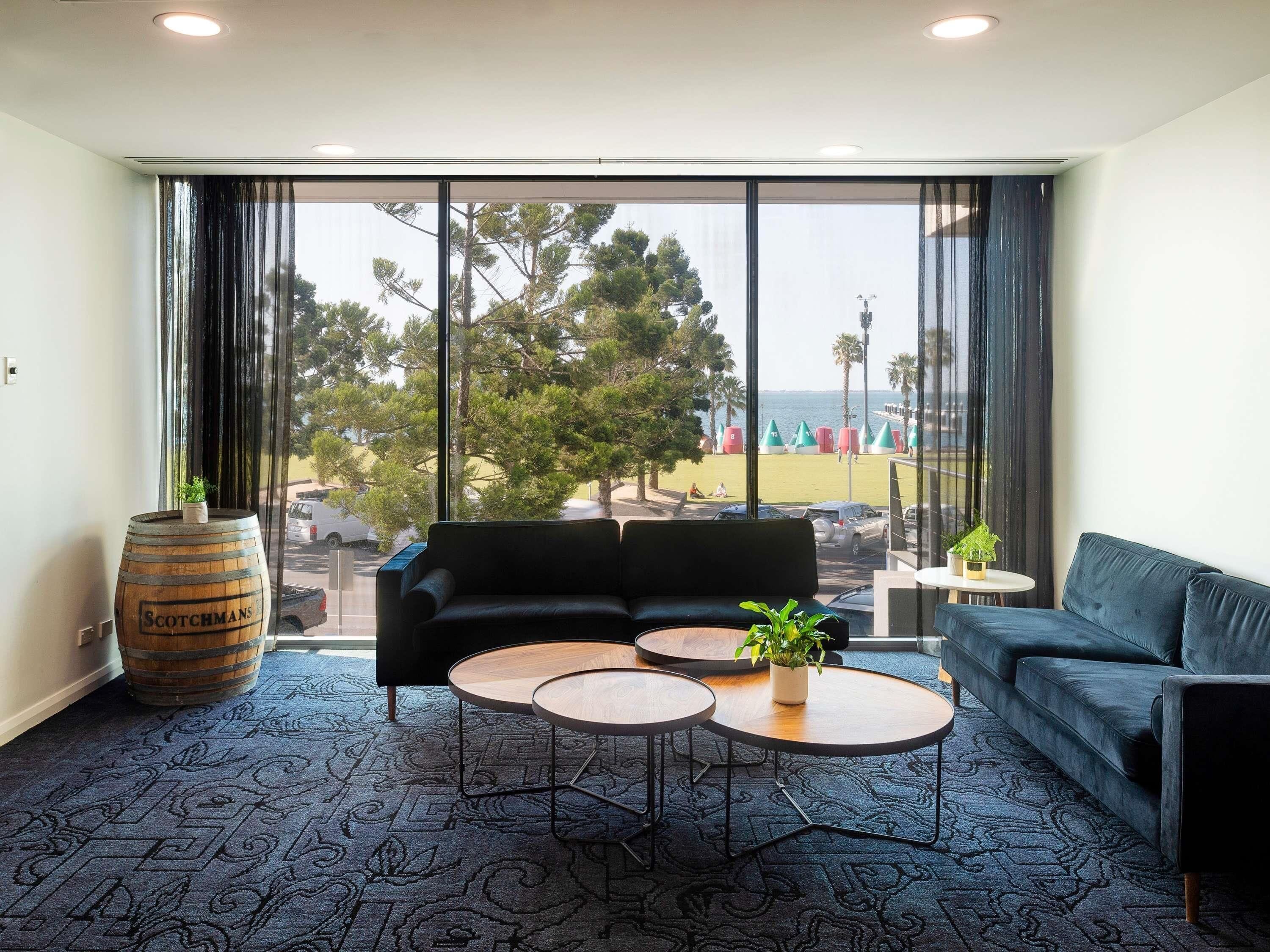 Una habitación moderna con un sofá, dos mesas redondas, un ventanal con vistas a una playa y árboles, y un barril etiquetado 'Scotchmans'.