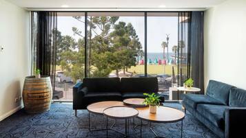Una habitación moderna con un sofá, dos mesas redondas, un ventanal con vistas a una playa y árboles, y un barril etiquetado 'Scotchmans'.