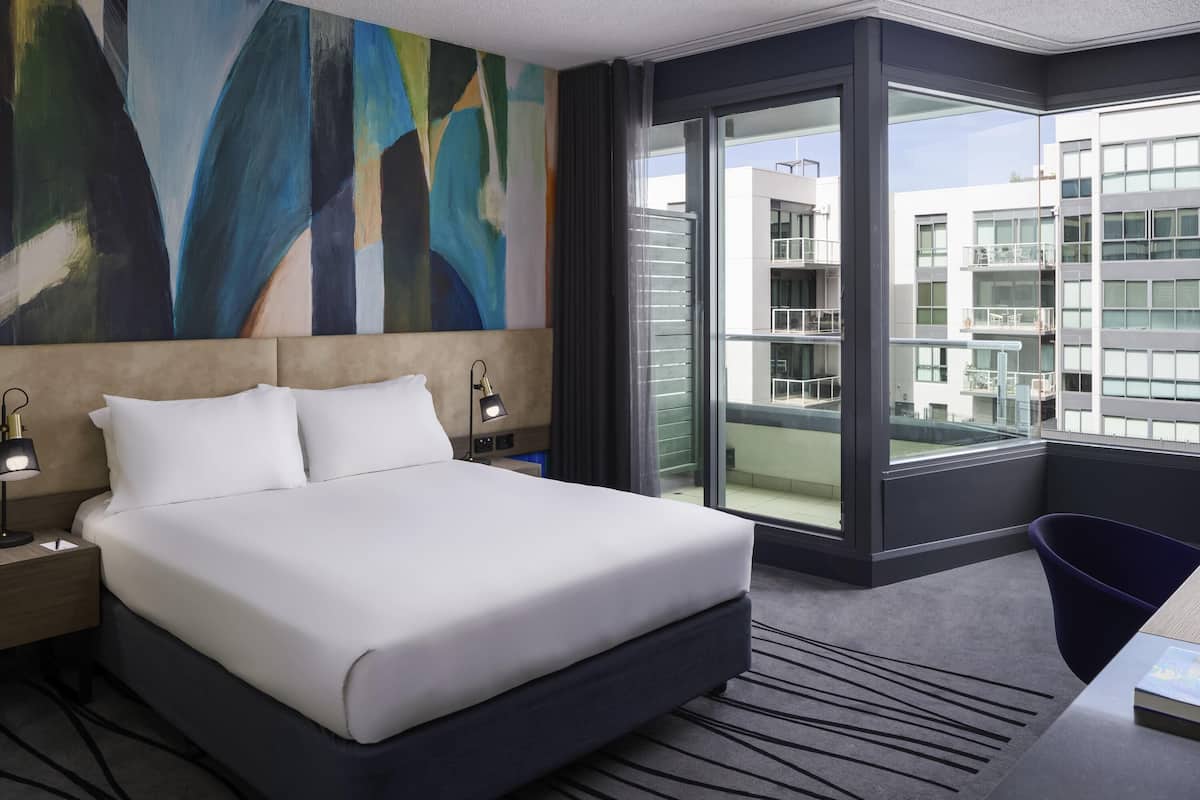 Habitación de hotel con una cama grande, un escritorio, una silla, una mesita de noche, una lámpara, un cuadro abstracto grande en la pared y vistas a un edificio a través de un ventanal amplio.