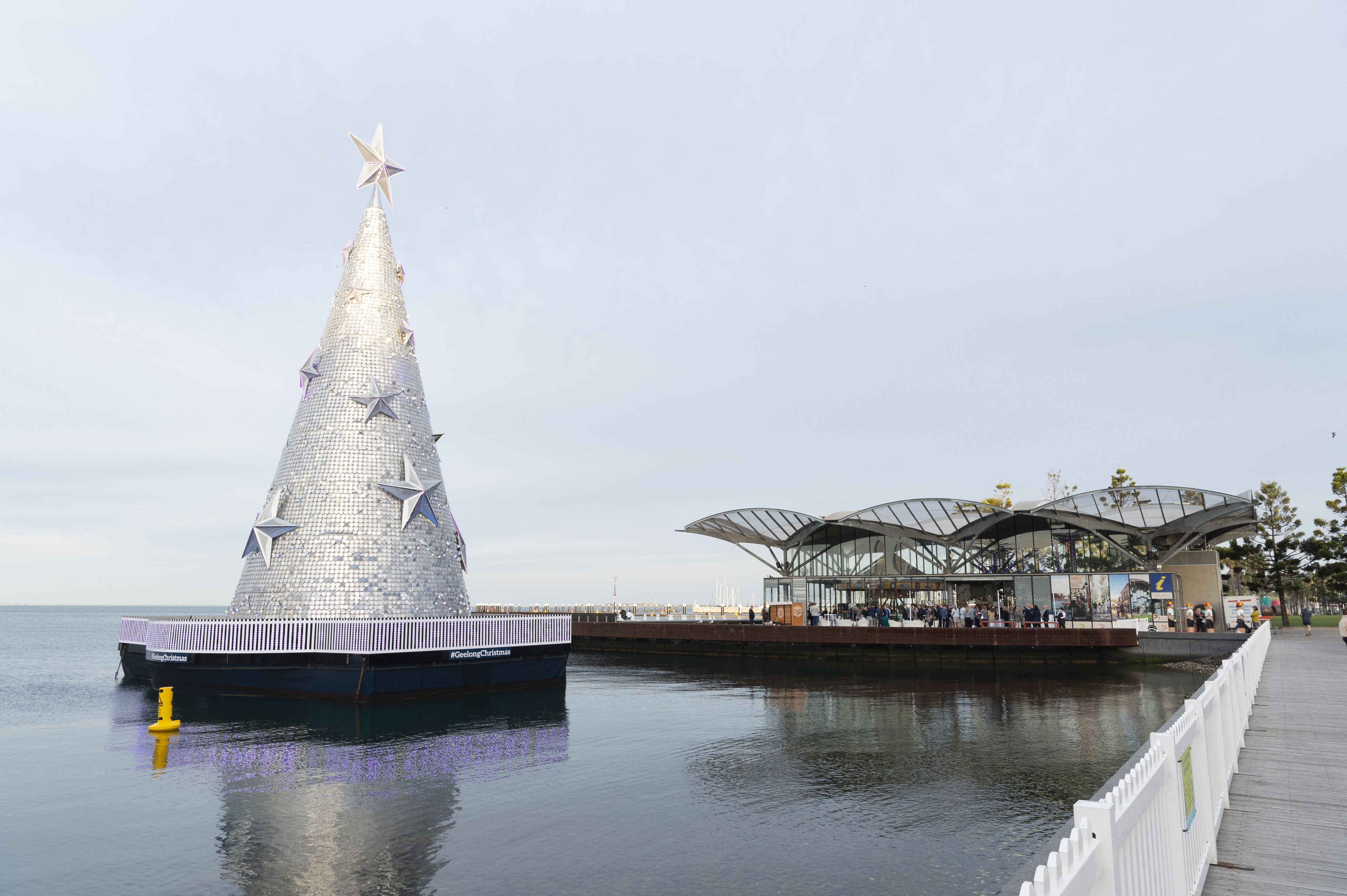 Una gran estructura metálica con forma de árbol de Navidad junto al agua, al lado de un moderno edificio de vidrio con techo curvo.