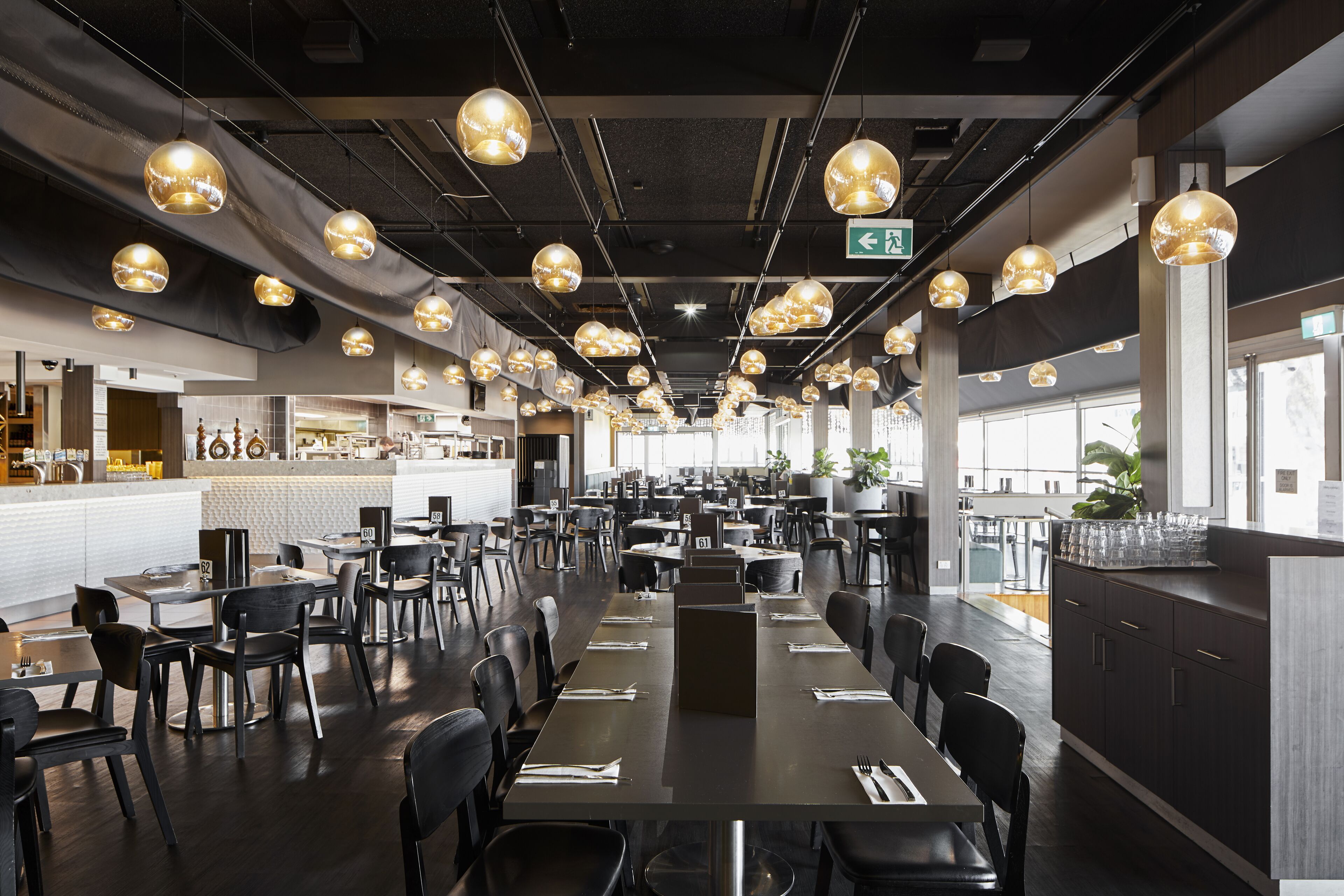 A spacious restaurant with multiple dining tables and chairs, pendant lighting, and a kitchen area in the background.