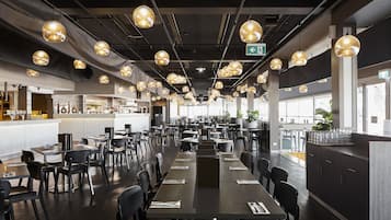 A spacious restaurant with multiple dining tables and chairs, pendant lighting, and a kitchen area in the background.
