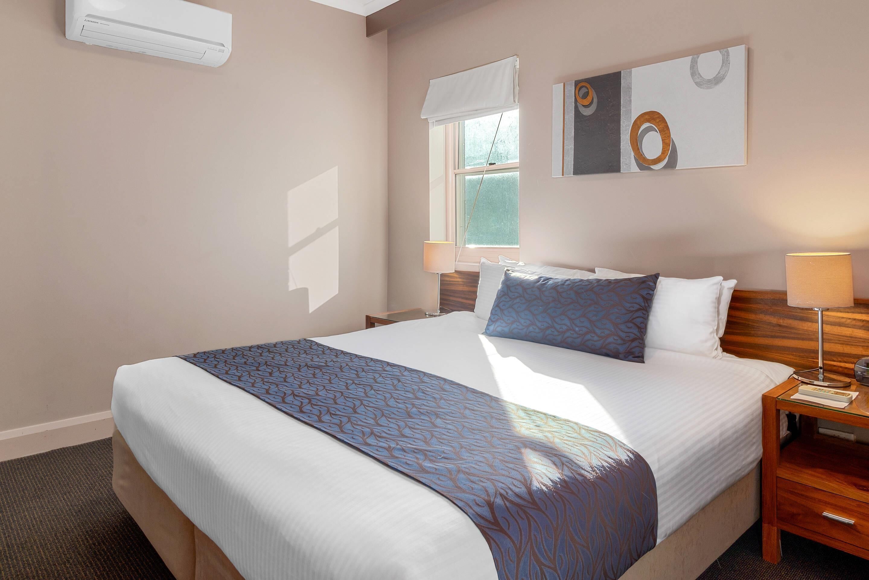 Una habitación de hotel con una cama grande, una mesita de noche con lámpara, una ventana con persianas y un aire acondicionado instalado en la pared.