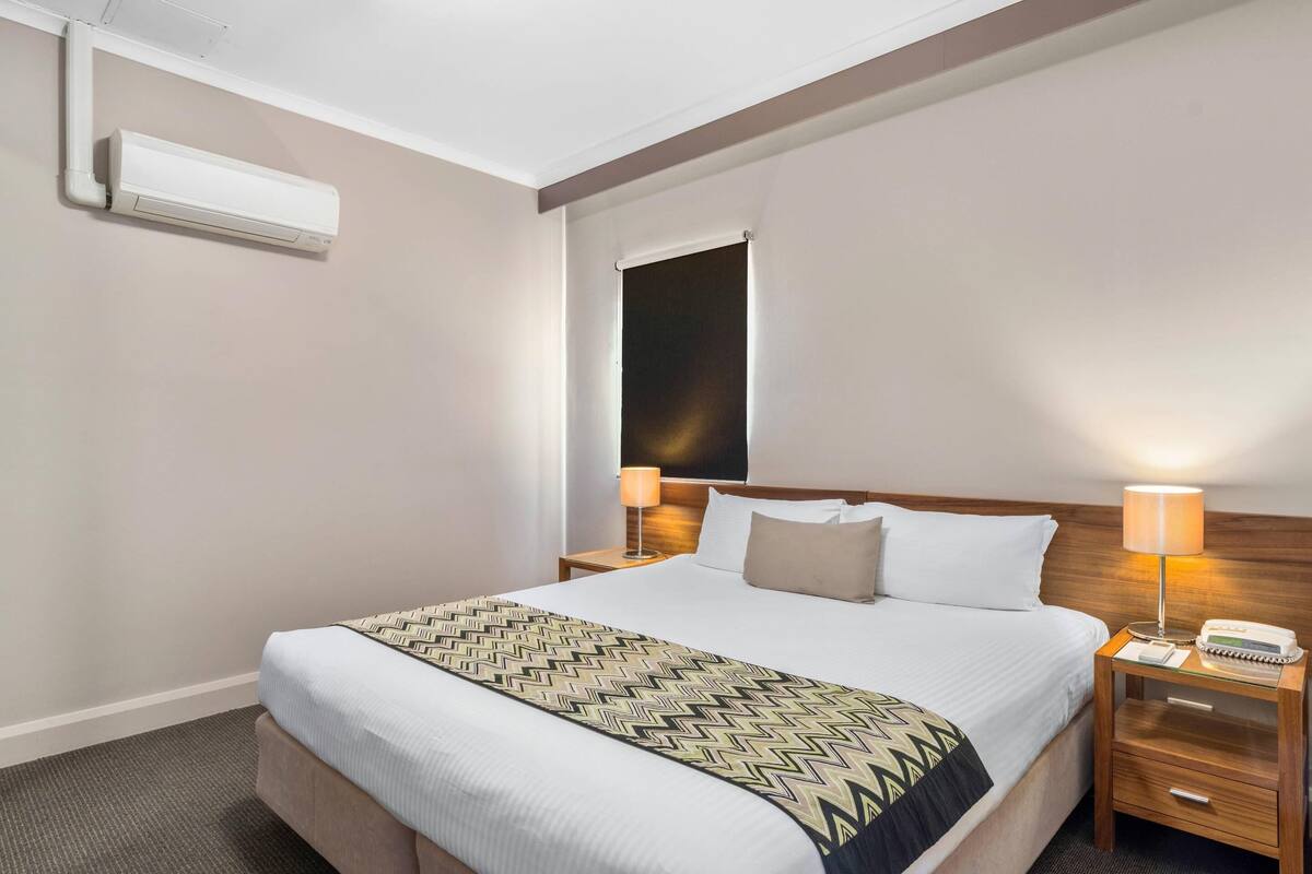 Una habitación de hotel con una cama, mesitas de noche, una lámpara y un aire acondicionado.