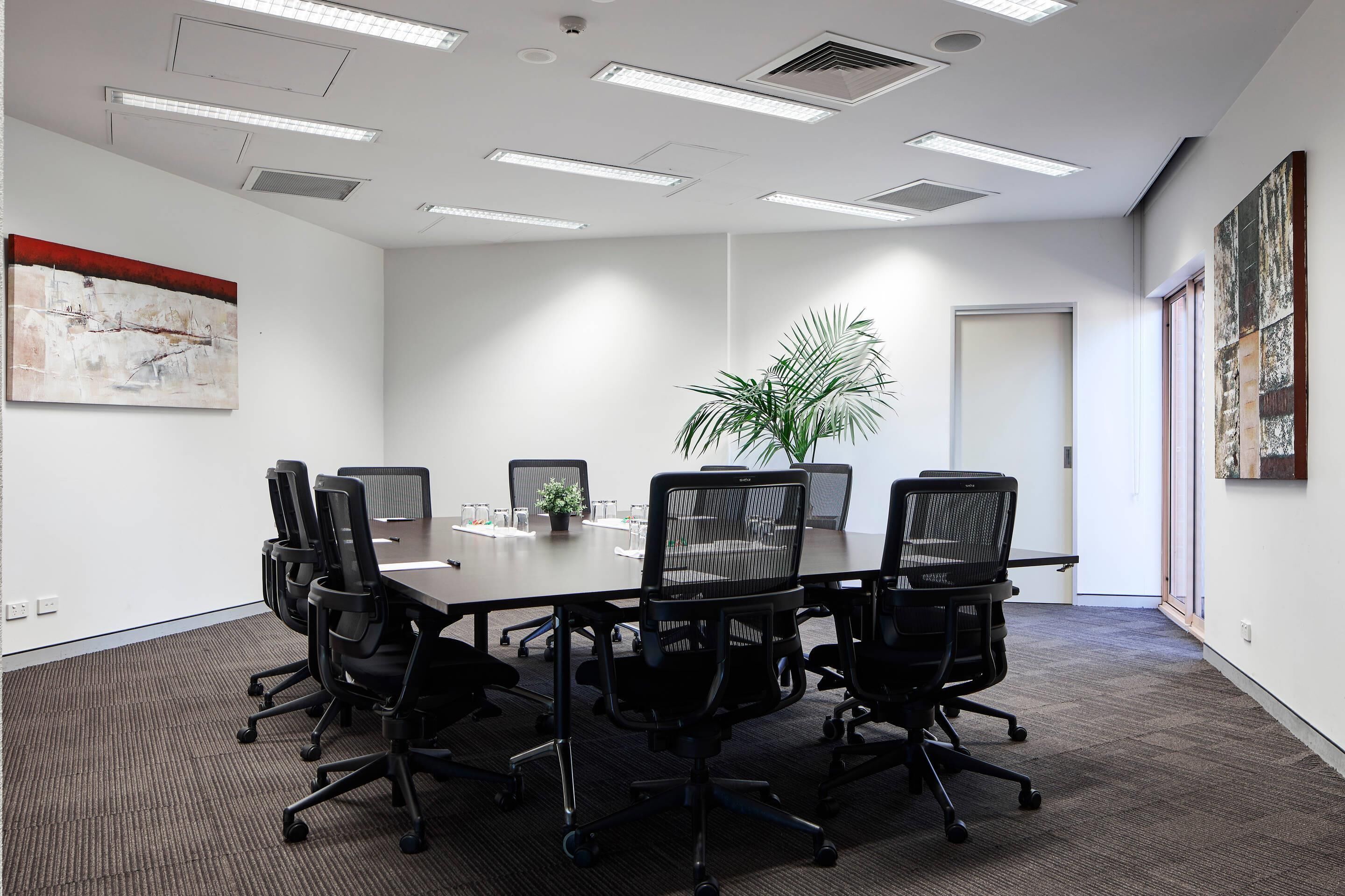 A conference room with a long table, black chairs, a potted plant, and artwork on the walls.