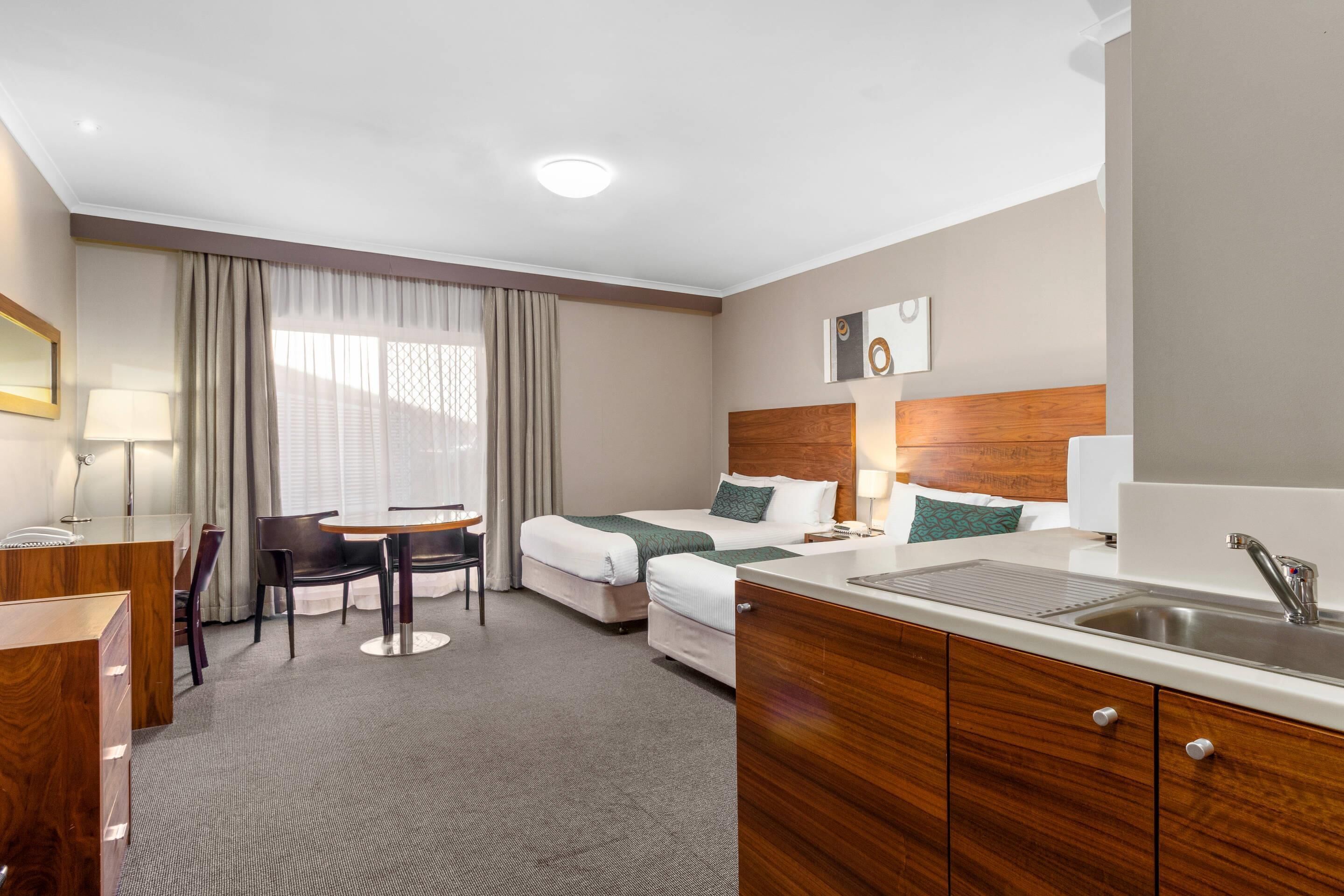 A hotel room with a bed, a desk, a chair, a small table, a sink, and a window with curtains.