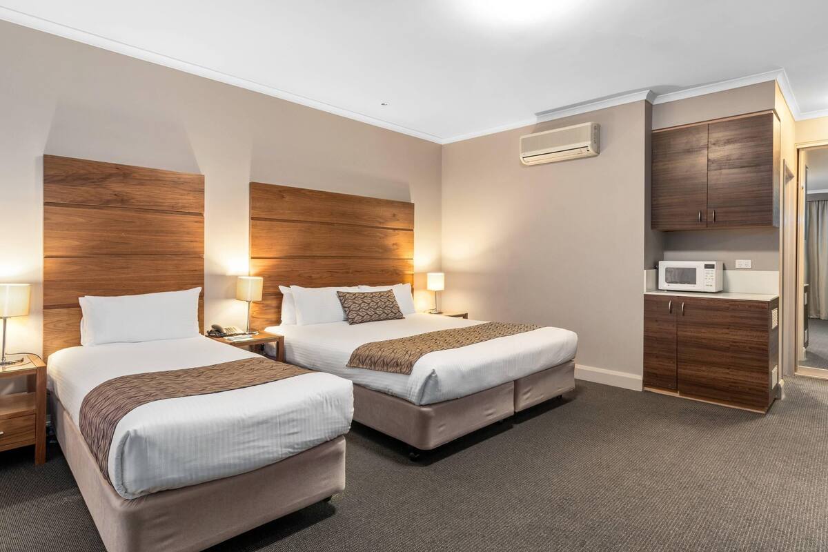 Una habitación de hotel con dos camas, un microondas, un armario y una mesita de noche con una lámpara.