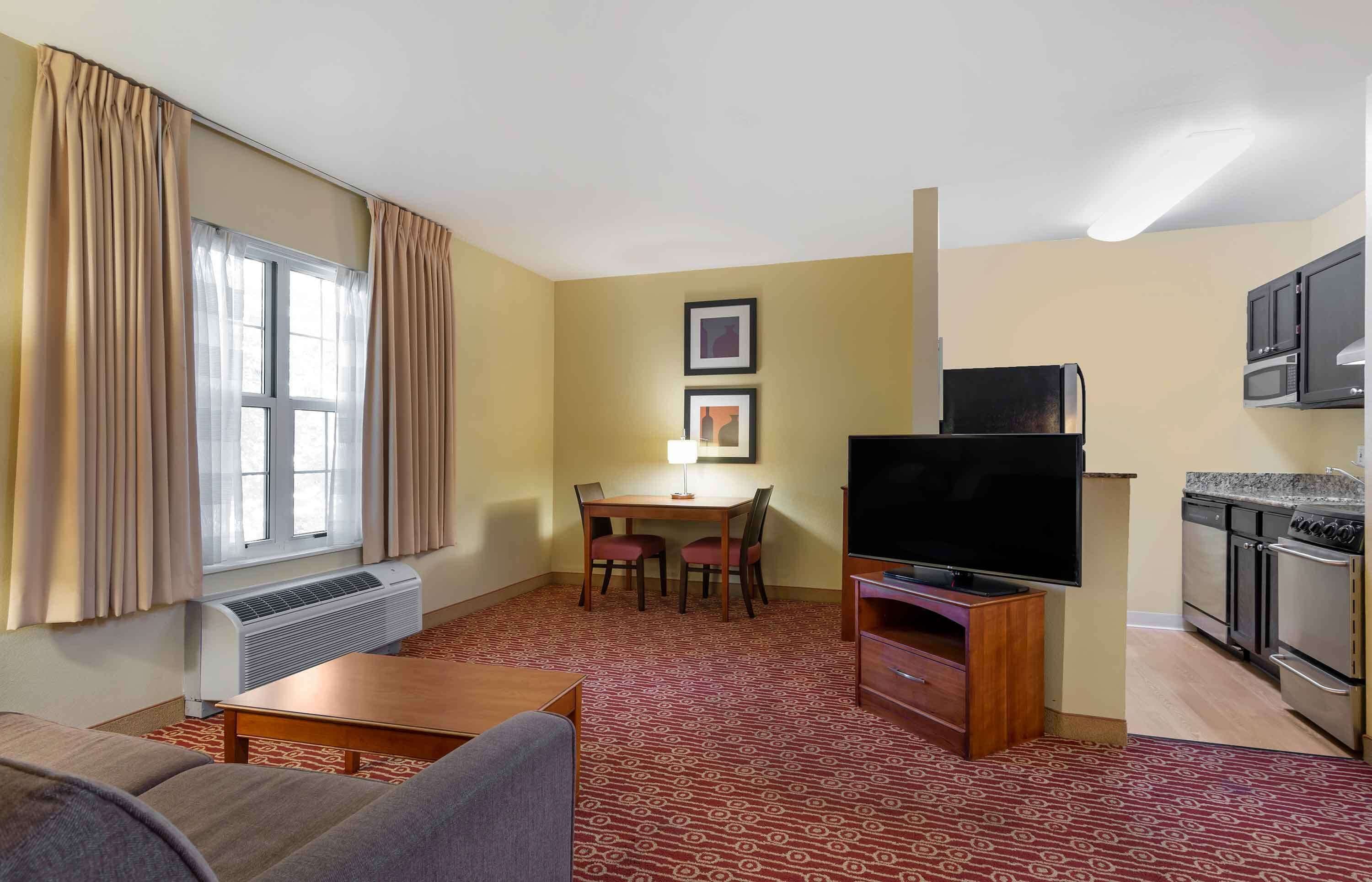 Habitación de hotel con sala de estar, una mesa de comedor con sillas, televisor de pantalla plana, kitchenette y ventana con cortinas.