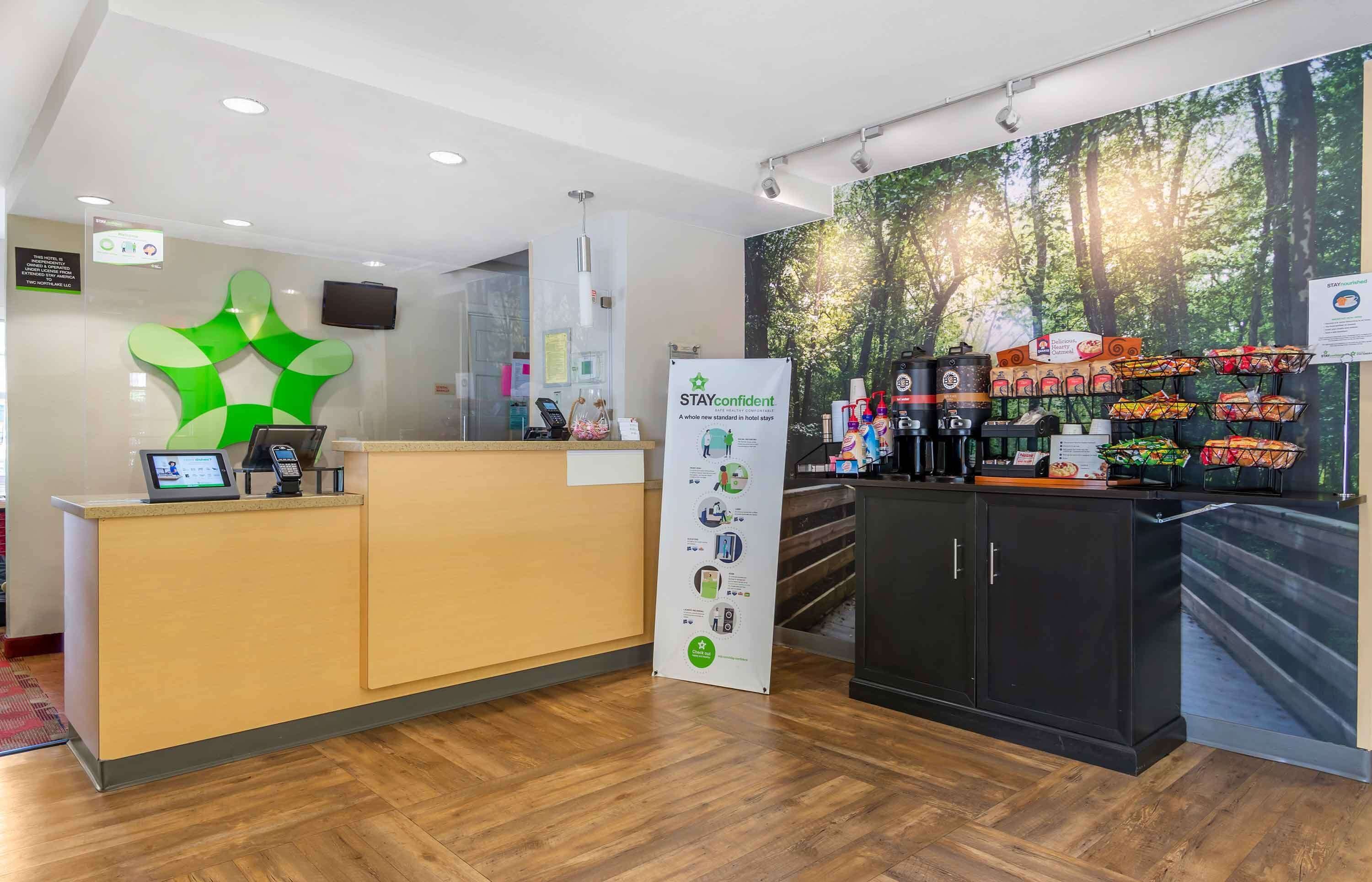 A hotel reception area with a modern design, including a reception desk, a large green abstract wall art, a poster titled "STAYCONFIDENT", and a coffee station with various items.
