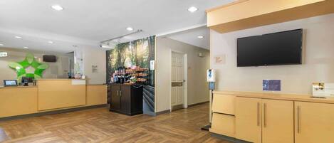 Una zona de recepción con piso de madera, un mostrador con caja registradora, un refrigerador y un televisor montado en la pared.