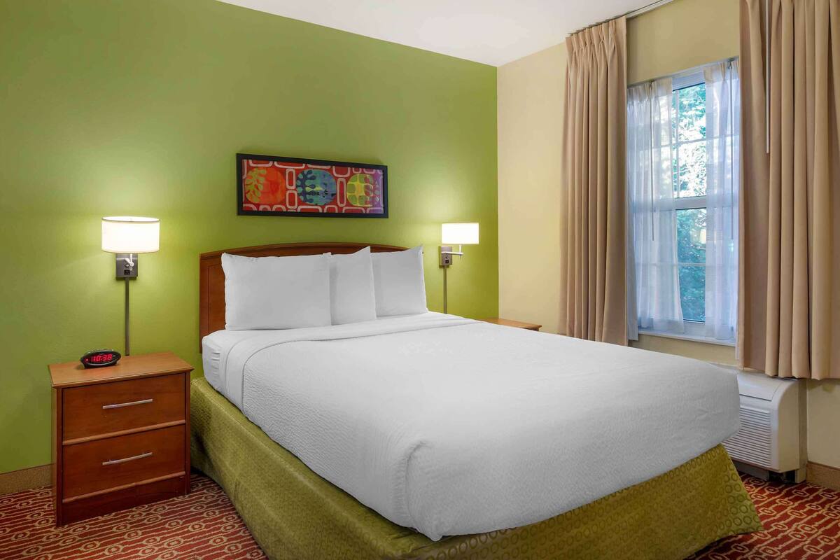 A hotel room with a bed, two bedside lamps, a nightstand with a clock, a window with curtains, and a colorful painting on the wall.
