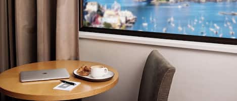 A room with a laptop, a cup of coffee, and croissants on a tray, with a view of a cityscape and a bridge through a window.