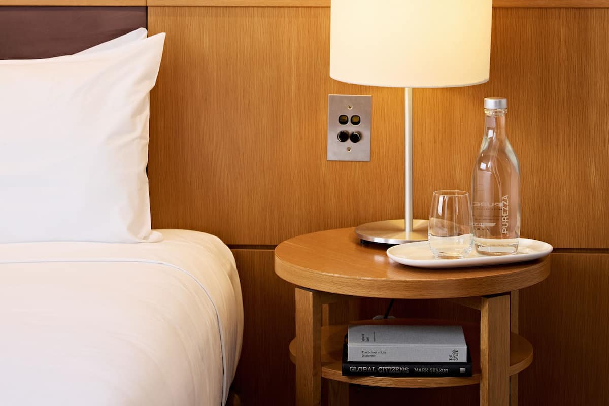 A hotel room with a striped wallpaper, a bed with white bedding, a bedside table with a lamp, a bottle, and a glass.