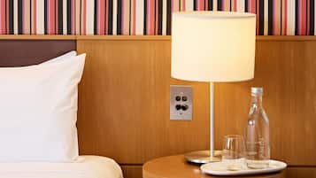 Hotelkamer met gestreept behang, een bed met wit beddengoed, een nachtkastje met een lamp, een fles en een glas.
