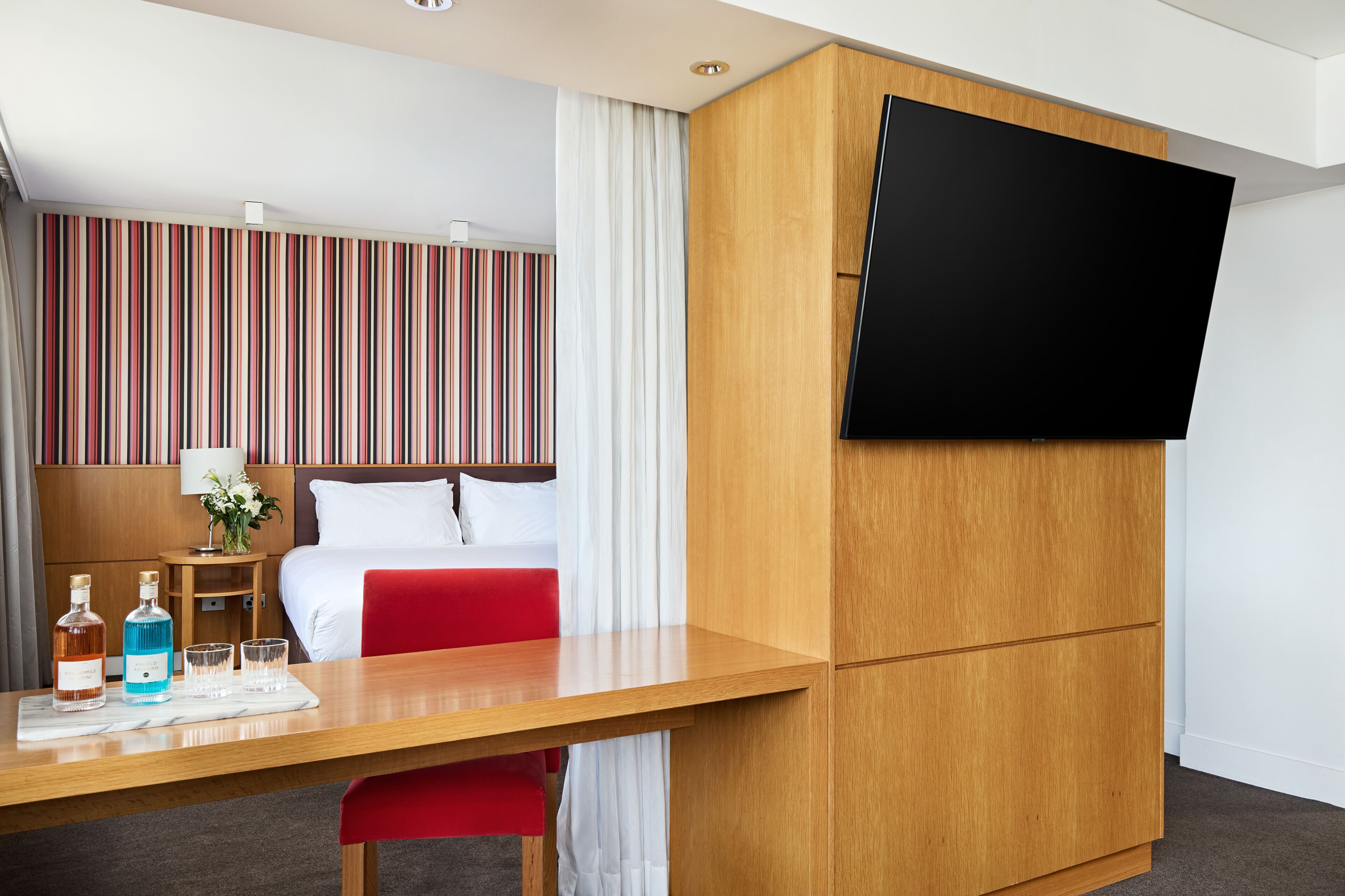 Une chambre d’hôtel avec un lit, un téléviseur fixé à un meuble en bois, un bureau avec une bouteille et des verres, et un mur à rayures.