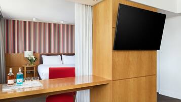 Une chambre d’hôtel avec un lit, un téléviseur fixé à un meuble en bois, un bureau avec une bouteille et des verres, et un mur à rayures.
