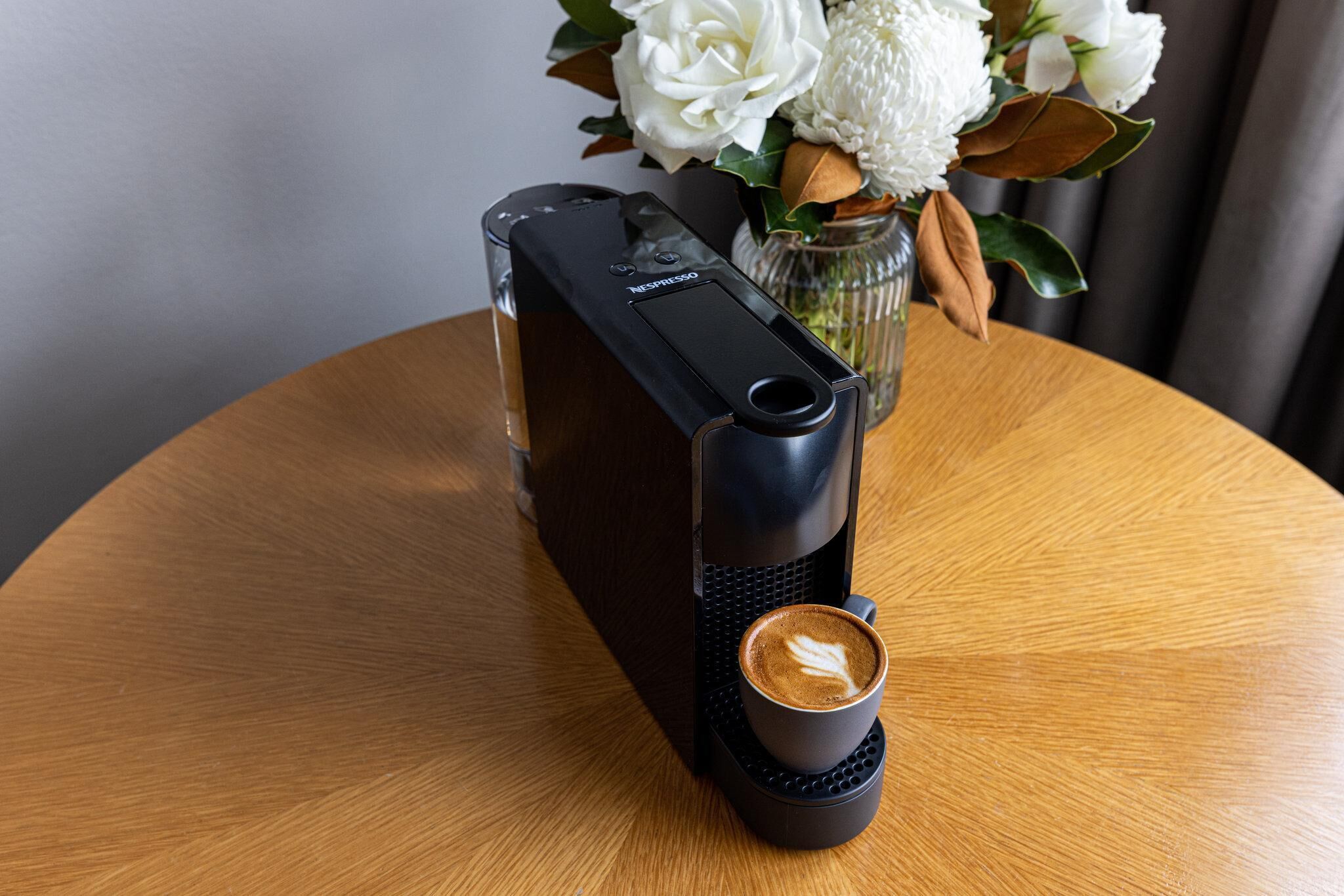 A Nespresso coffee machine on a wooden table with a cup of coffee and a vase of flowers in the background.