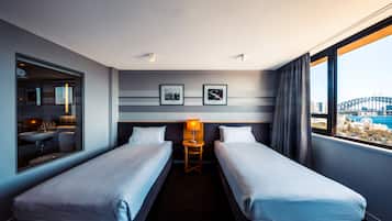 A hotel room with two beds, a small table with a lamp, a window with a view of a bridge, and framed pictures on the wall.