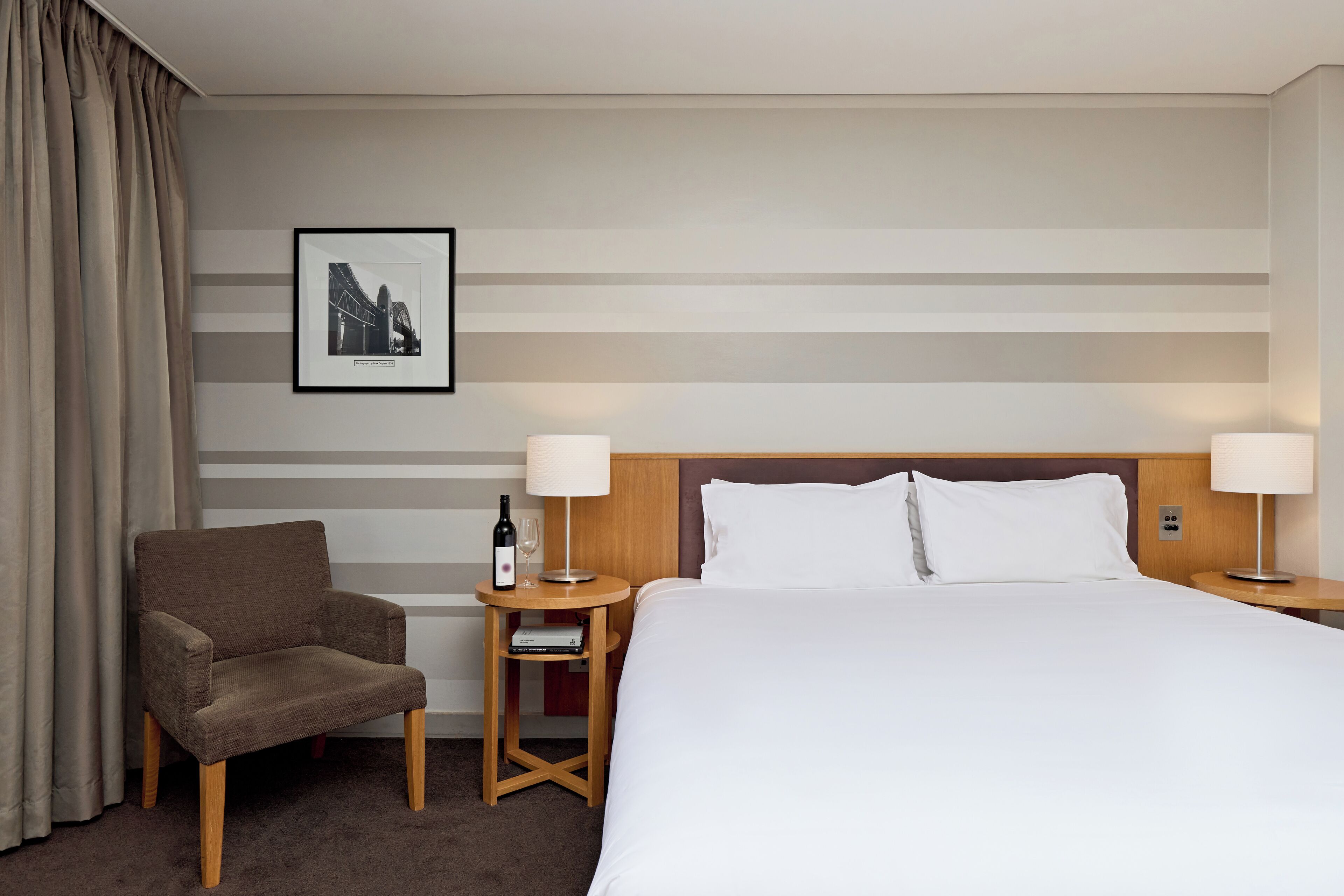 Habitación de hotel con cama, dos mesitas de noche, una silla, un cuadro en la pared y una botella de vino sobre una de las mesitas.