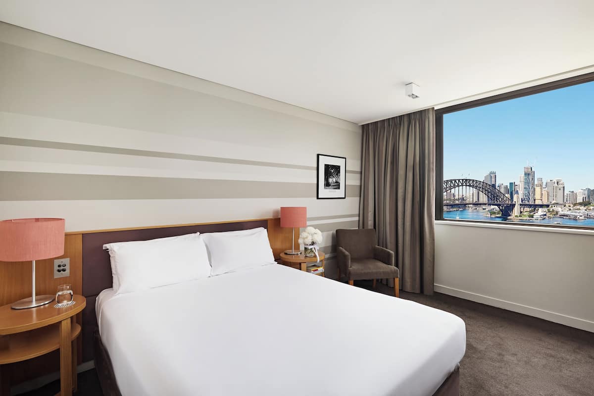 A hotel room with a large bed, two bedside tables, a chair, a window with a cityscape view, and a framed picture on the wall.