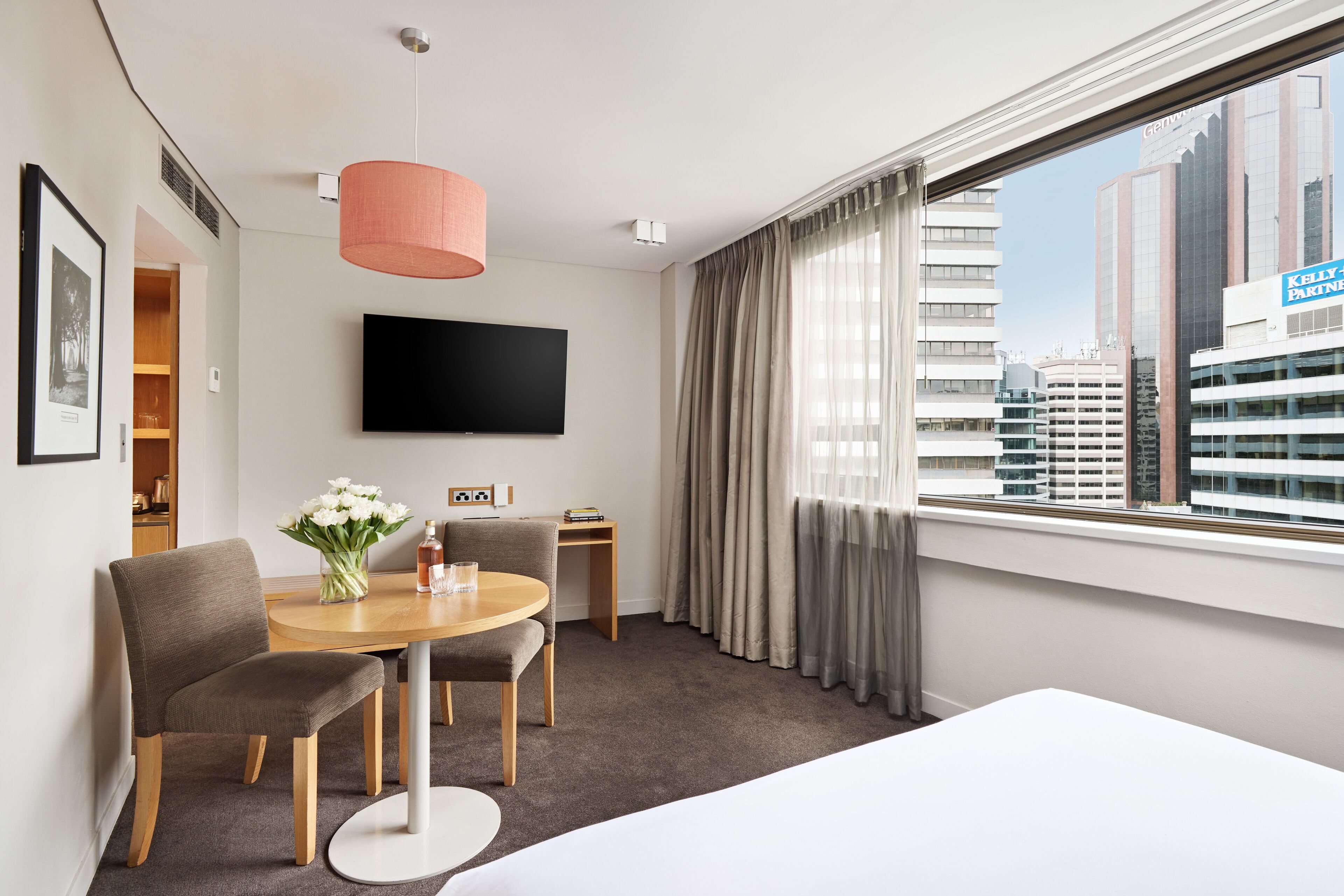 Habitación de hotel con cama, televisor montado en la pared, mesa de comedor con sillas, mesita auxiliar y ventana con cortinas.
