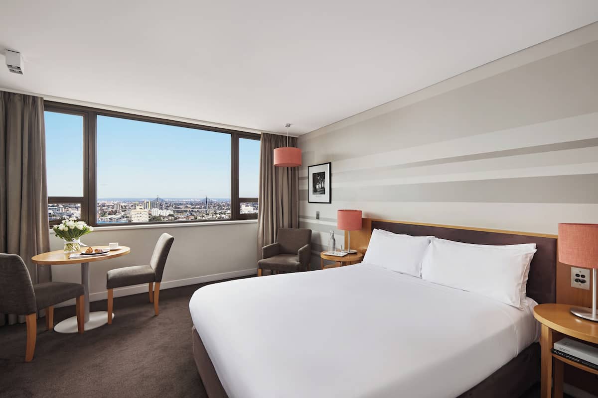A hotel room with a large bed, a desk with a chair, a small table with flowers, a chair, a nightstand with a lamp, and a view of the city through large windows.
