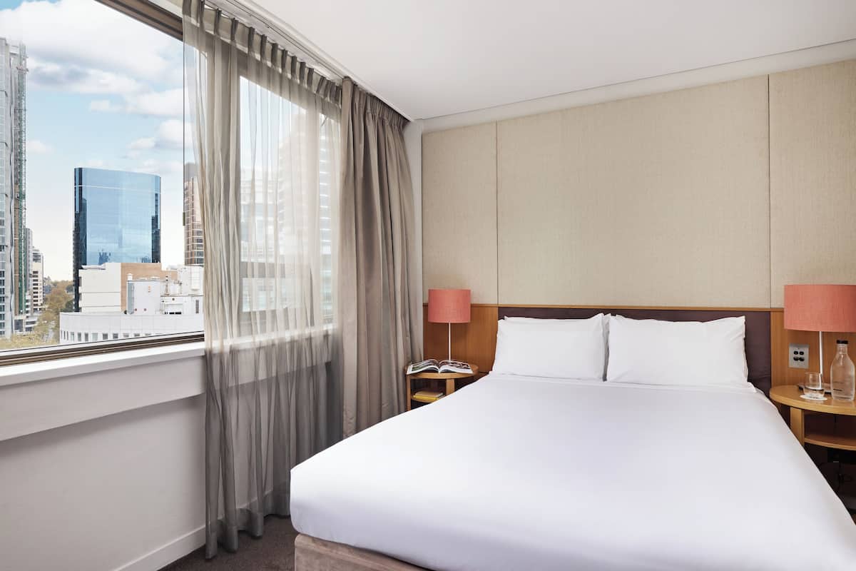 A hotel room with a large bed, a nightstand with a lamp, and a view of a cityscape through a window with sheer curtains.