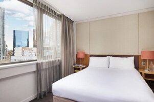 A hotel room with a large bed, a nightstand with a lamp, and a view of a cityscape through a window with sheer curtains.