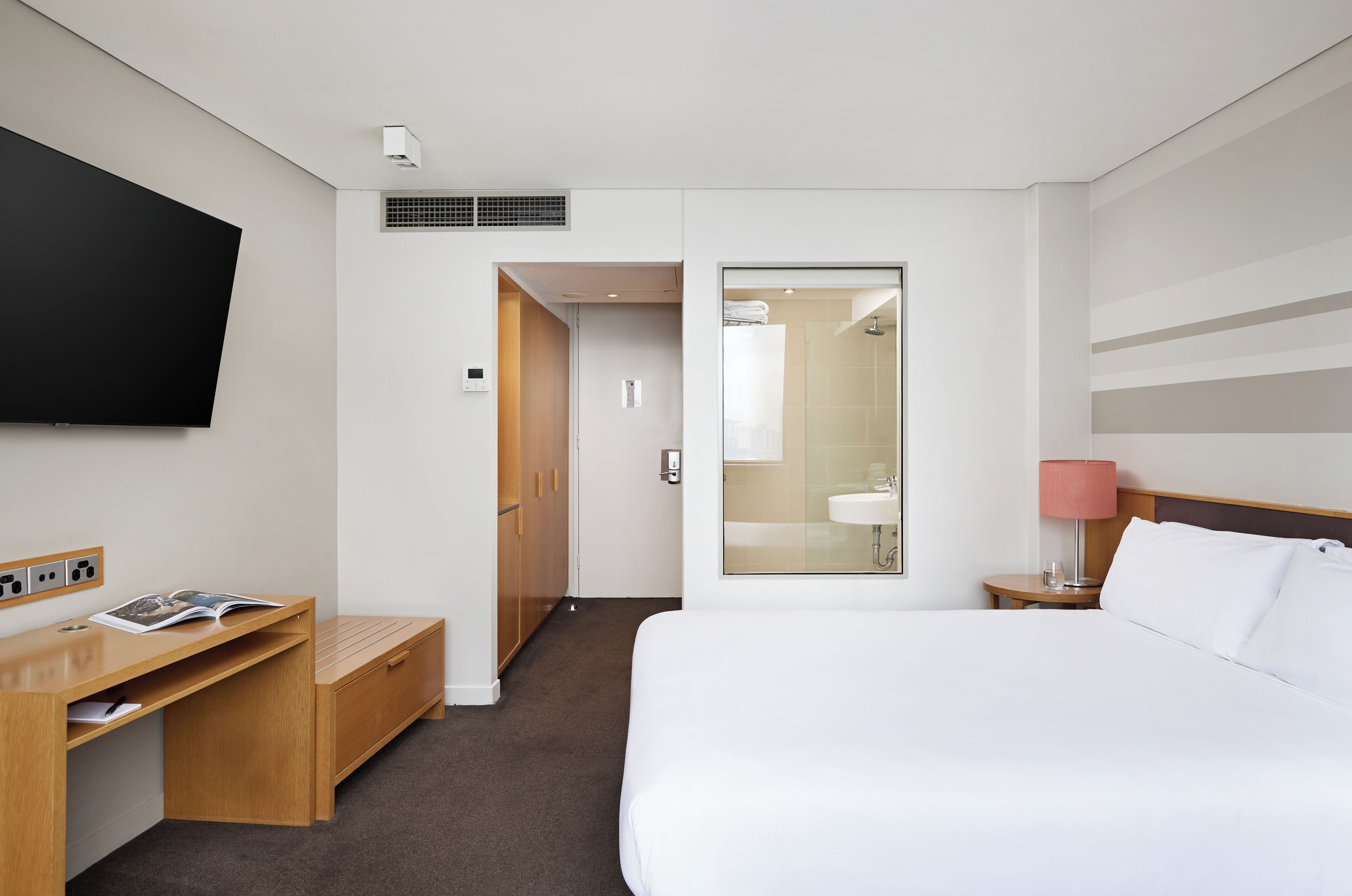 A hotel room with a bed, a desk with a magazine, a TV mounted on the wall, a bathroom with a shower, and a bedside lamp.