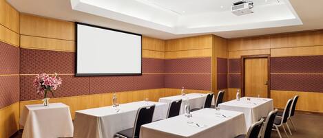 Un salón de conferencias con mesas, sillas, una pantalla de proyector y un arreglo floral.