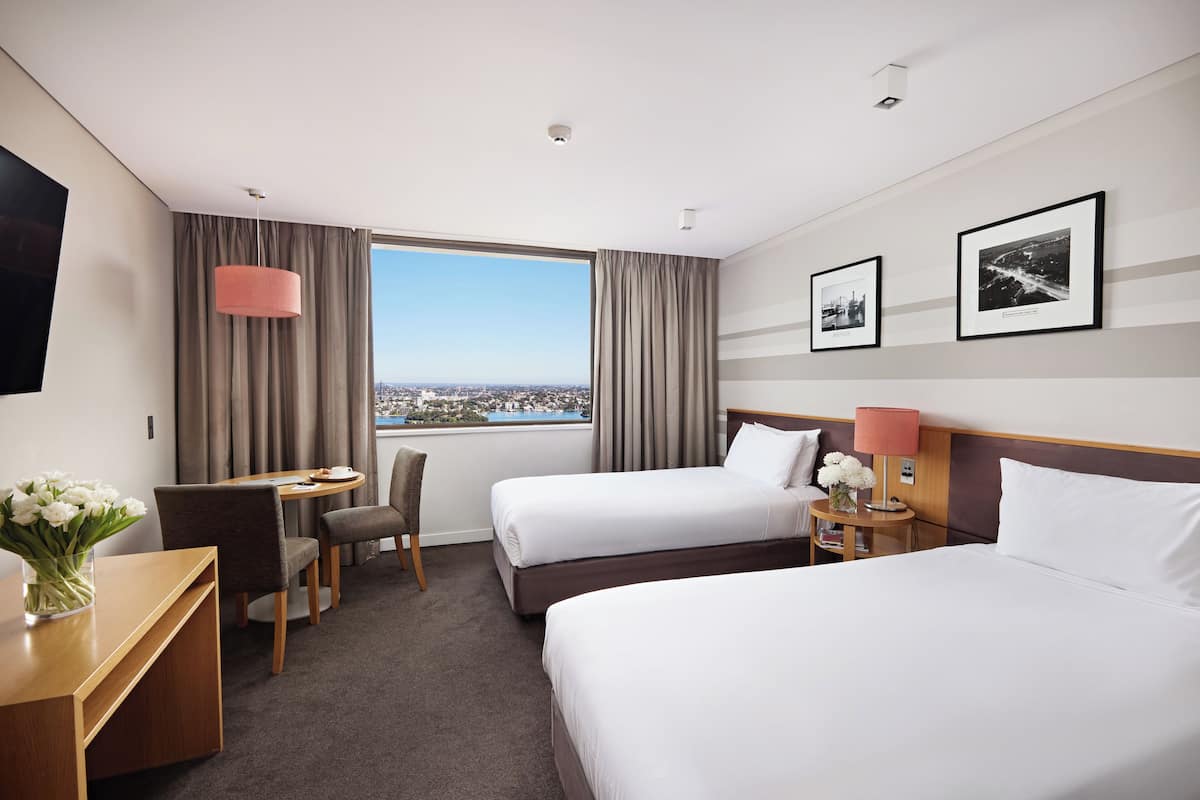 A hotel room with two beds, a desk, a chair, a small table with a lamp, and a view of the city.