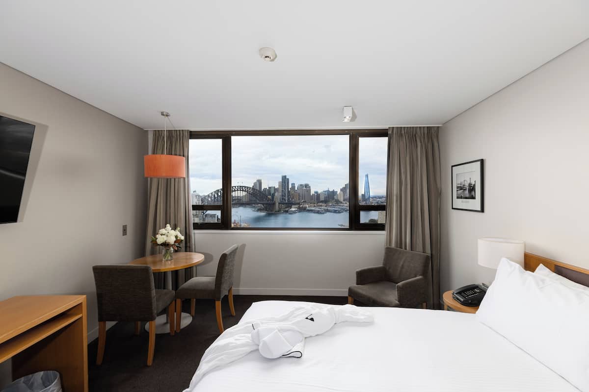 A hotel room with a bed, a desk, a chair, a small table with a vase of flowers, and a view of a cityscape through large windows.
