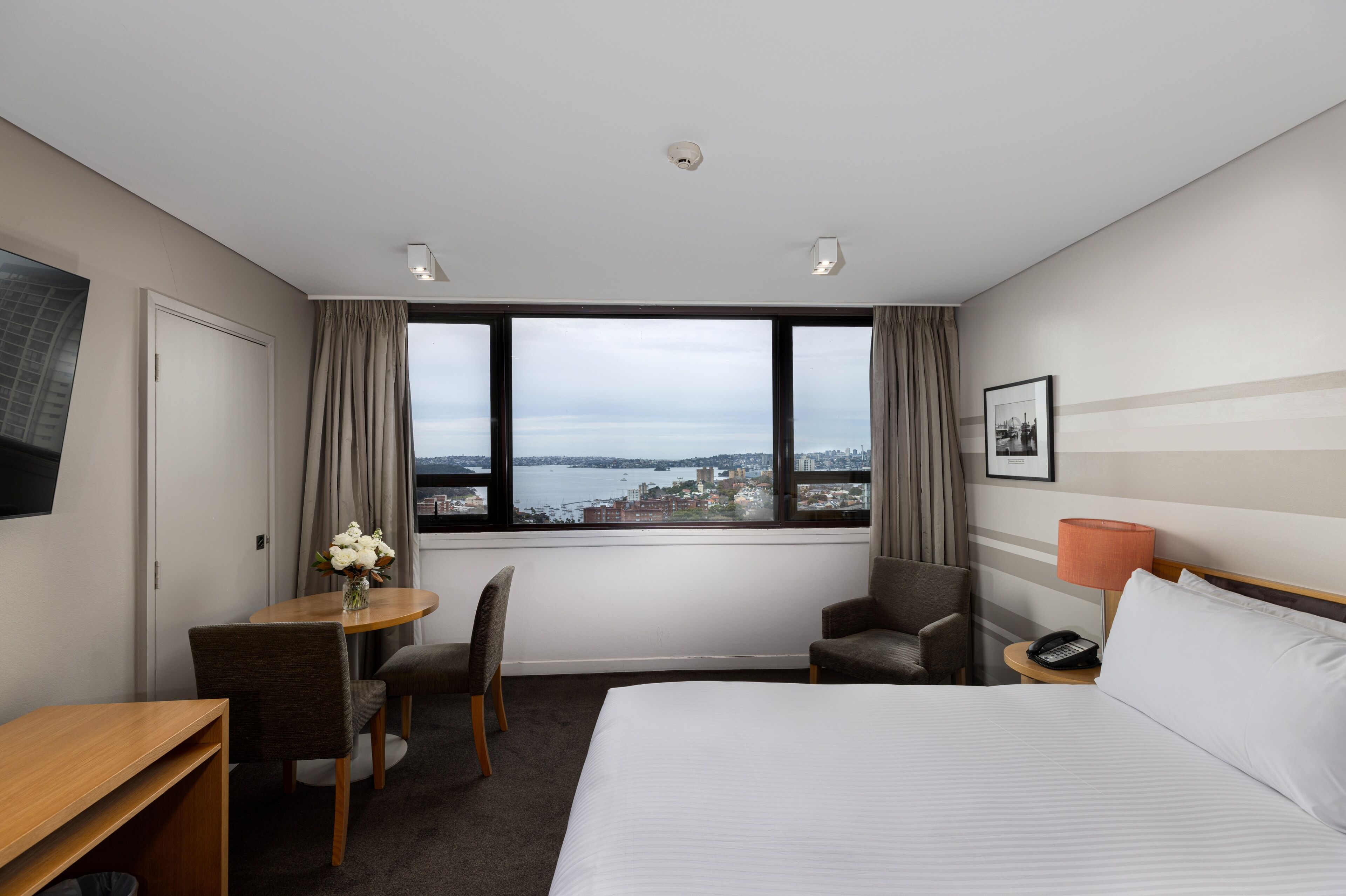 A hotel room with a large bed, a desk with a chair, a small table with a vase of flowers, a chair, a nightstand with a lamp, and a view of a body of water and buildings.