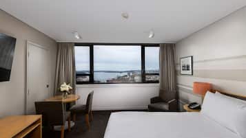 A hotel room with a large bed, a desk with a chair, a small table with a vase of flowers, a chair, a nightstand with a lamp, and a view of a body of water and buildings.