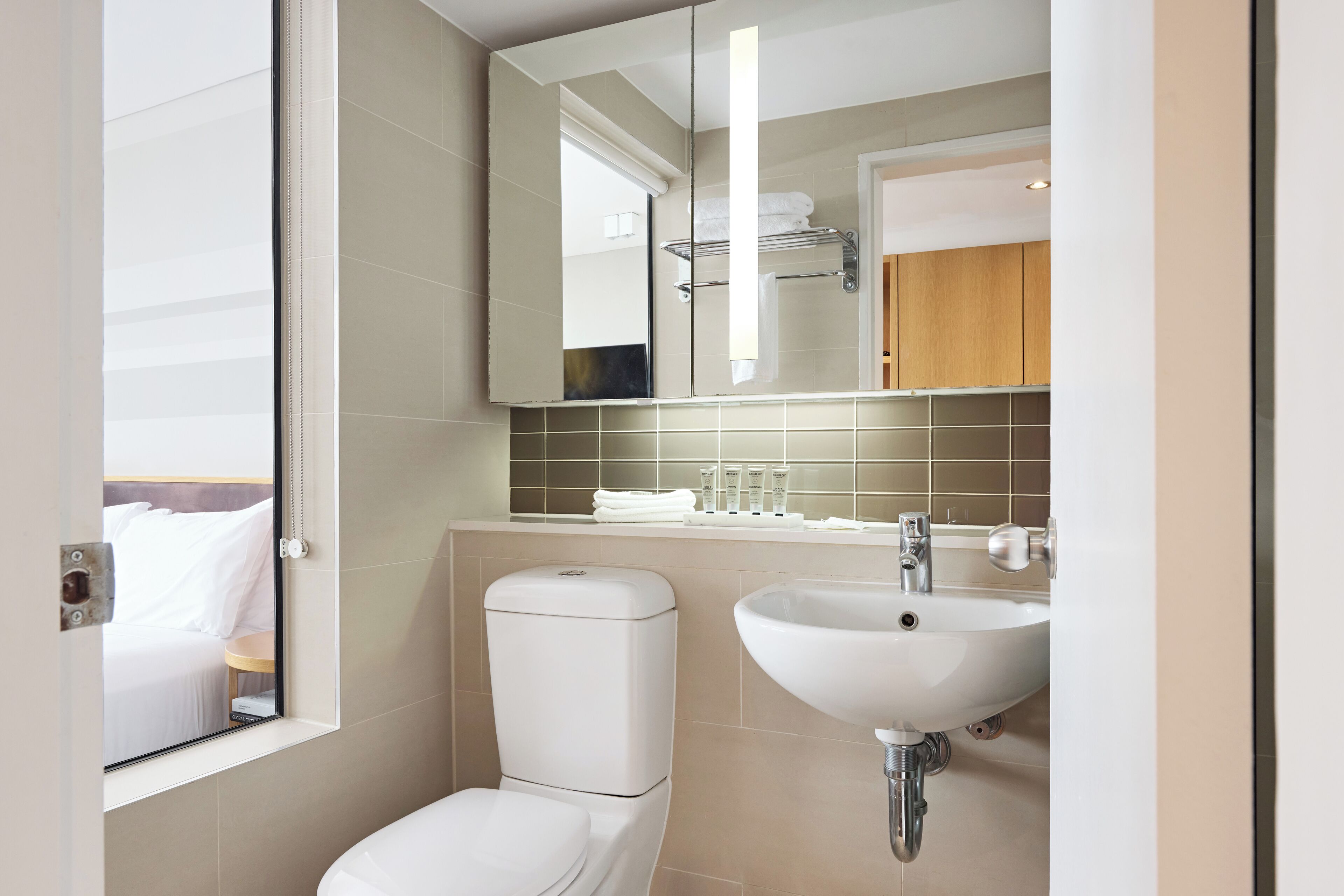 A modern bathroom with a toilet, sink, and a mirror reflecting a bed and a towel rack.