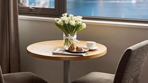 Ein Hotelzimmer mit einem runden Tisch, zwei Stühlen, einer Blumenvase, einer Tasse und einem Teller mit einem Croissant. Ein großes Fenster bietet Blick auf eine Stadtsilhouette und eine Brücke.
